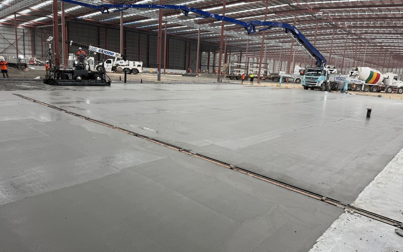 A laser screed and a Lotos boom pump working a freshly placed slab inside a steel-framed warehouse