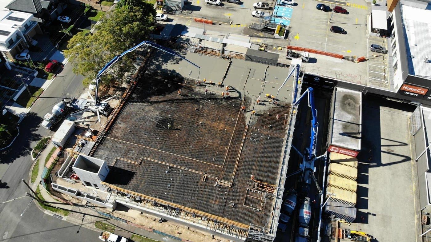 Aerial view of a reinforced ground slab mid-pour on a constrained site, with two boom pumps and agitator trucks working off the street