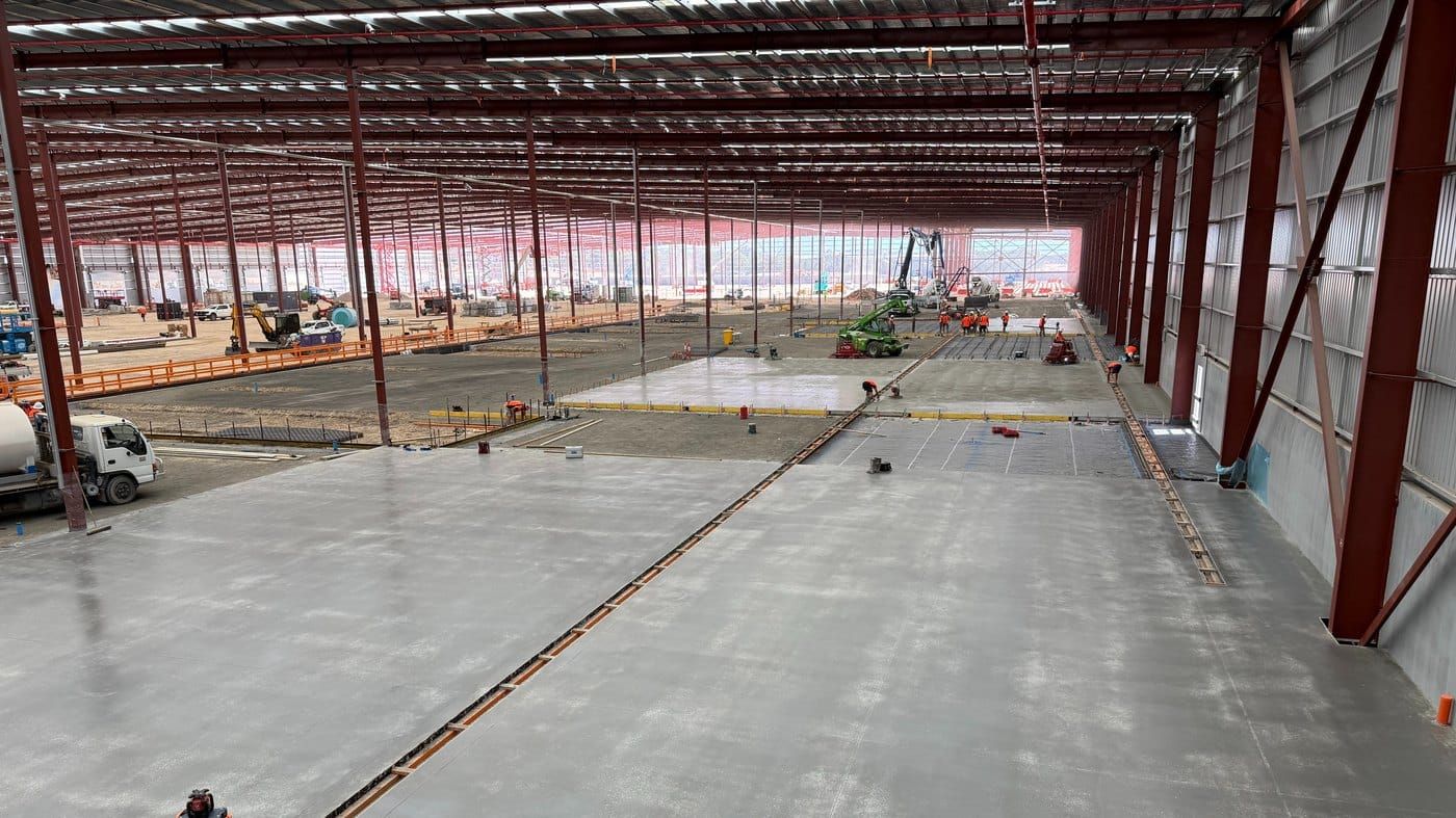 A large warehouse floor being placed and finished inside a red steel portal frame, with the screed crew and a telehandler at work